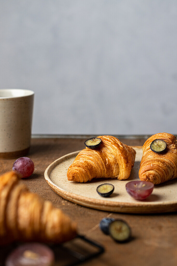 Tasty fresh baked croissants served on plate with fruits placed near cup of tea on wooden table in morning time in light room