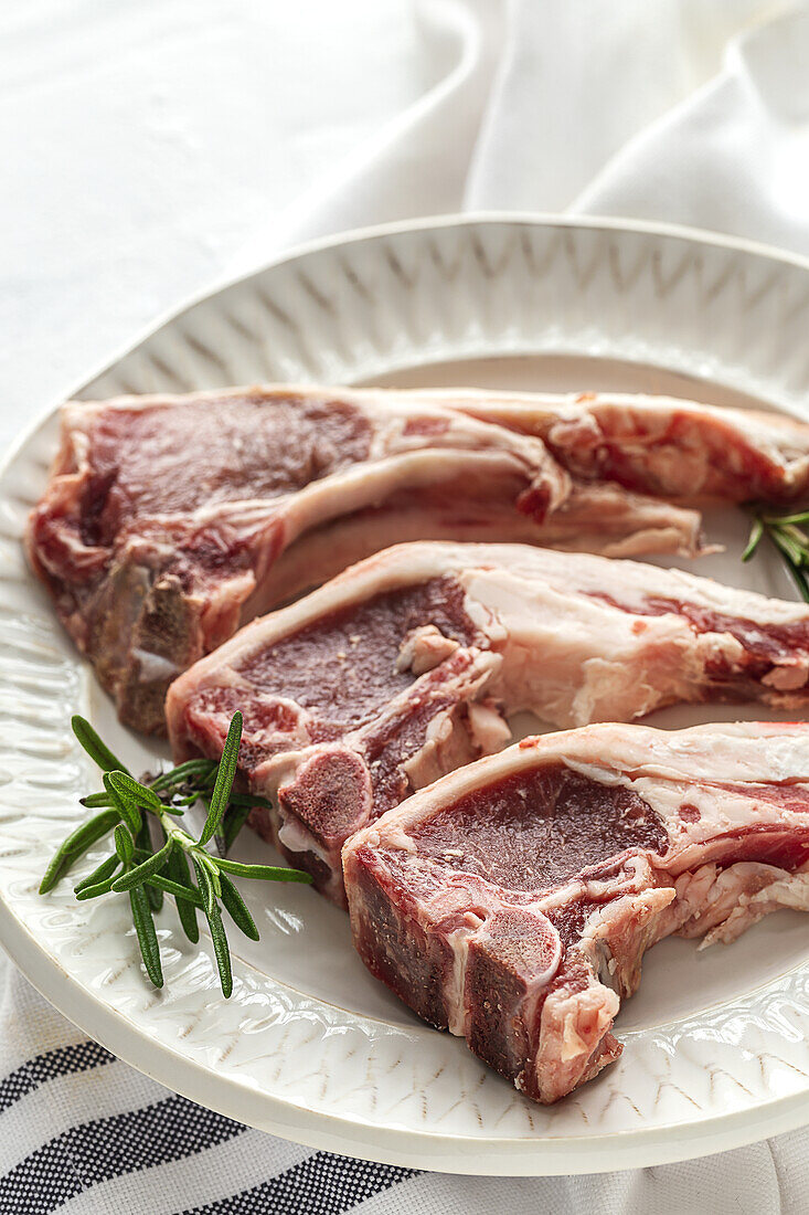 From above of uncooked lamb chops served on white ceramic plate with fresh green rosemary on table in light kitchen