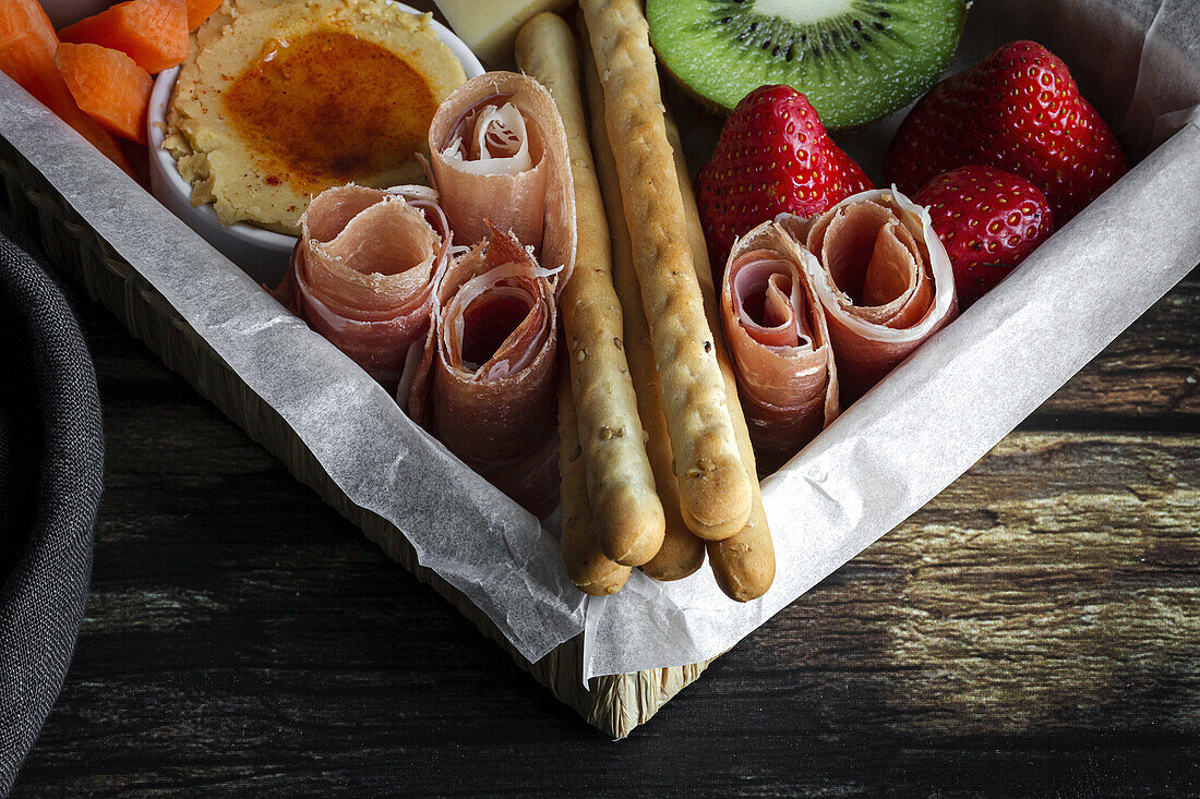 Von oben Brunch-Box mit verschiedenen Wurst- und Käsesorten und Knäckebrot in der Nähe von reifen Kiwis, süßen Erdbeeren und geschälten Mandarinen in der Nähe von Marmelade in einem Glas auf einem Holztisch in der Nähe einer Serviette