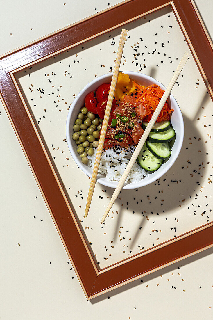 From above white bowl with tasty poke dish and chopsticks placed behind frame on table covered with sesame seeds