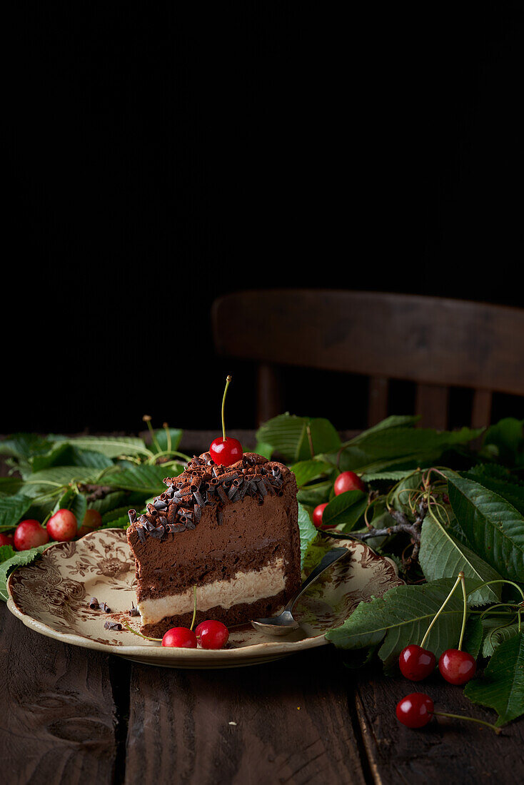 Appetitanregender hausgemachter Kuchen mit Schokoladen- und Vanillemousse auf einer Platte und mit frischen Kirschen dekoriert neben einem Haufen grüner Blätter auf einem Holztisch