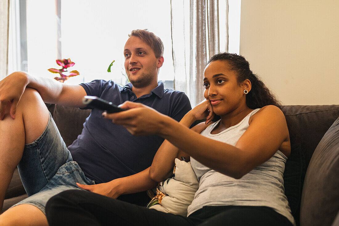Delighted multiracial couple switching channels with remote control while relaxing and watching TV