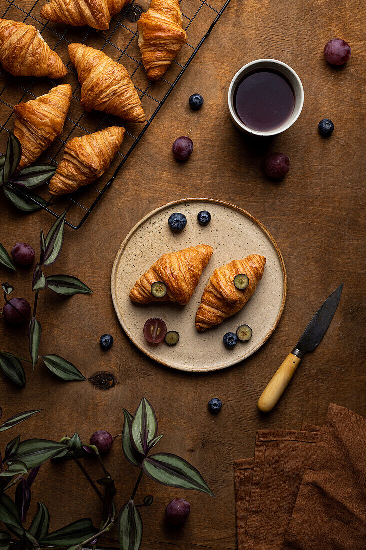 From above of tasty fresh baked croissants served on plate with fruits placed near cup of tea on wooden table in morning time in light room