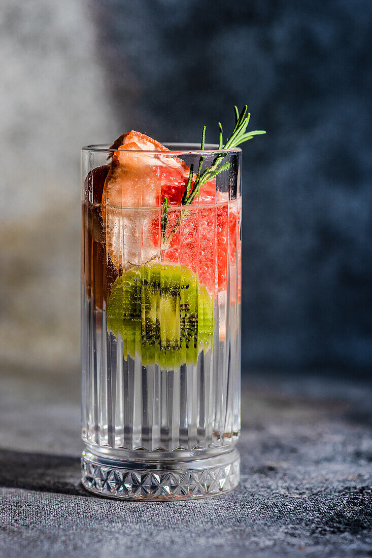 Tropischer Sommercocktail aus Wodka und Sprite mit reifen Früchten und Rosmarin auf grauem Hintergrund