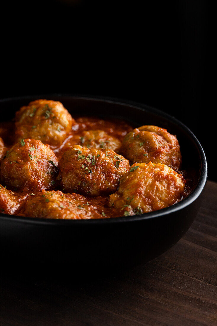 From above of black bowl with delicious meatballs in tomato sauce with herbs placed on wooden board in dark room