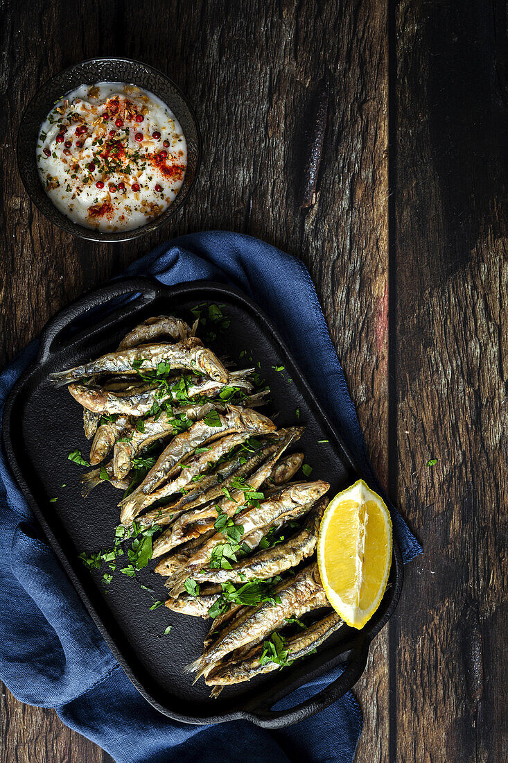 Top view of tasty fried anchovies with juicy lemon piece and chopped parsley on tray against savory sauce