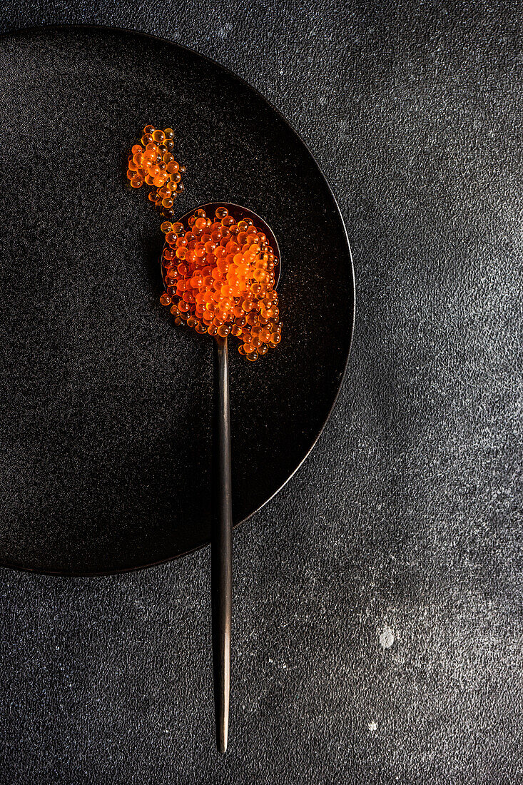 Schwarzer Löffel voll mit rotem Kaviar auf schwarzem Teller und gleichfarbigem Stein-Beton-Tischhintergrund