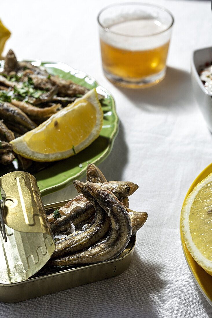 From above of delicious fried anchovies served on plate and can with lemon and placed on white table with glass of beer
