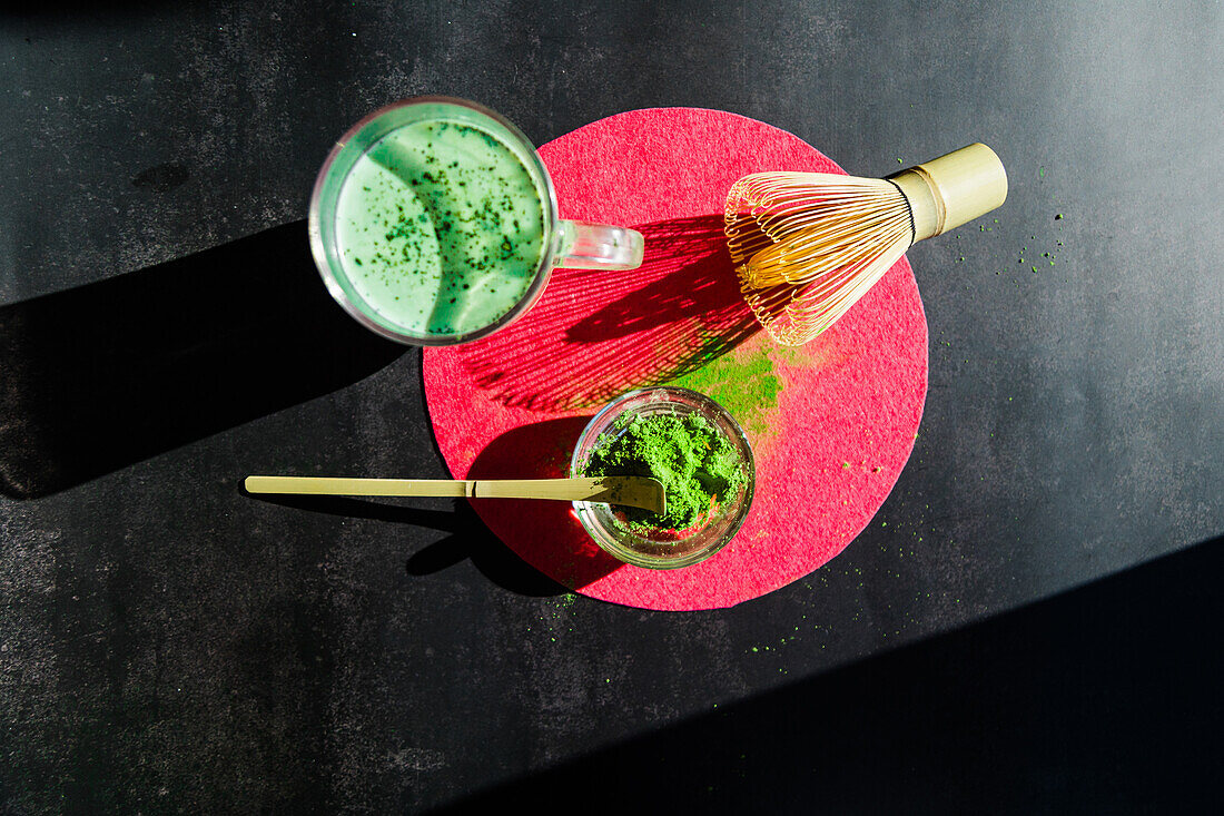 Top view of healthy matcha tea and dried powder near wooden chasen and chashaku on bright round stand on black table at sunlight