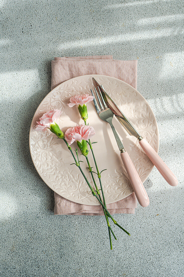 Von oben gedeckter Tisch mit Teller und Serviette neben rosa Nelkenblüten auf Betontisch