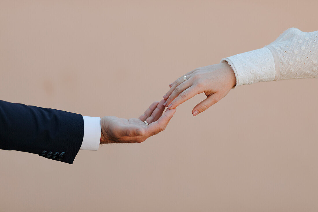 Anonymes romantisches Brautpaar in eleganter, stilvoller Hochzeitskleidung mit Verlobungsringen berührt sanft die Hände vor einem beigen Hintergrund