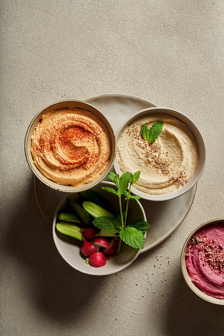 Top view of bowls with assorted hummus served on table with fresh cucumbers and radish