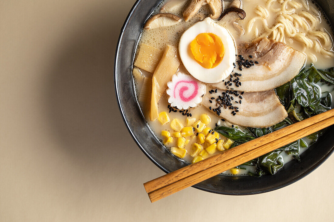 Top view of appetizing Japanese ramen with boiled egg and mushrooms served in bowl with wooden chopsticks against beige background