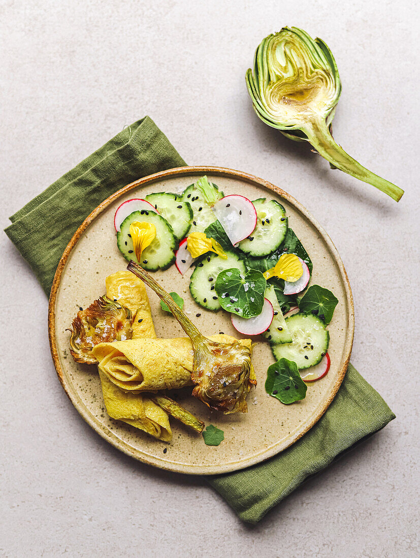 Top view of plate with omelette roll and radish and cucumber salad with herbs placed near artichoke and napkin on gray table
