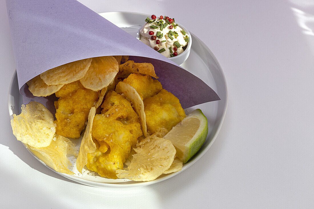 Appetizing fried chips and fish in paper wrapping served on plate with sauce on table with bright sunlight in cafe