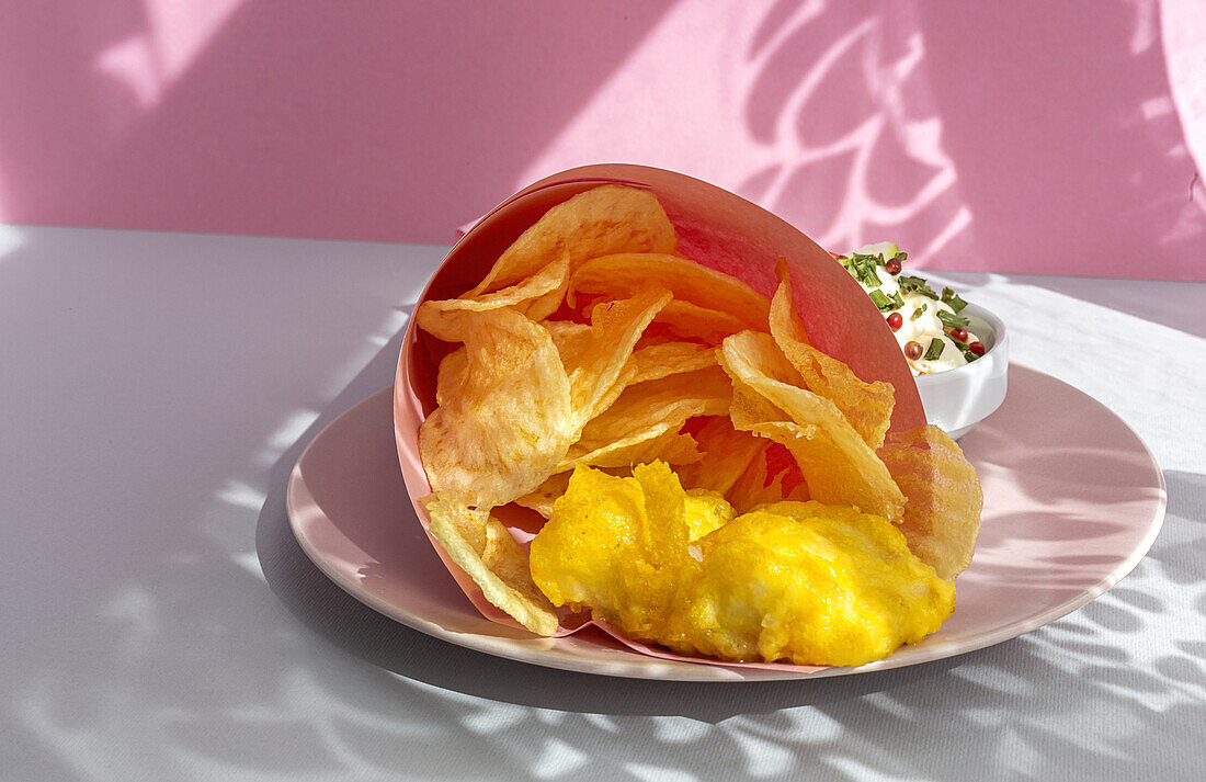 Appetitlich gebratene Pommes frites und Fisch in Papierverpackung auf einem Teller mit Soße auf einem Tisch mit hellem Sonnenlicht in einem Café serviert