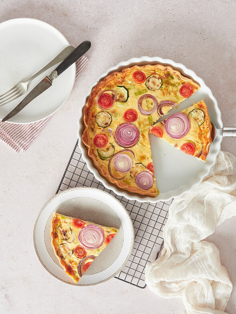 Top view of tasty vegetable quiche served on plate and baking pan near napkin and grating on table in kitchen