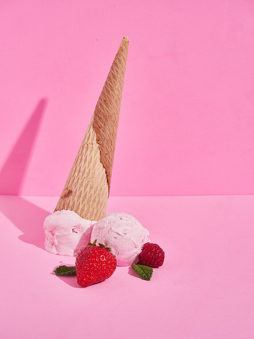 Köstliche süße Erdbeereis-Kugeln mit heruntergefallenen knusprigen Kegeln in der Nähe von reifen Beeren vor rosa Hintergrund mit Schatten im Studio