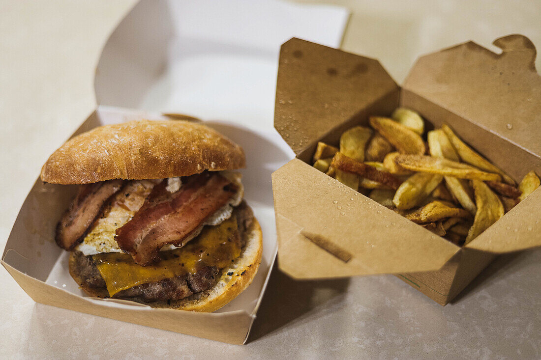 High angle of appetizing hamburger with bacon and crispy potato wedges served on table in carton packages