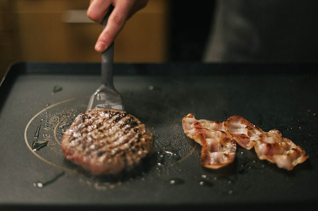 Anonymer Koch mit Schulterblatt, der auf einem schwarzen Herd appetitlich gegrillte Frikadellen für Burger und Speckscheiben zubereitet