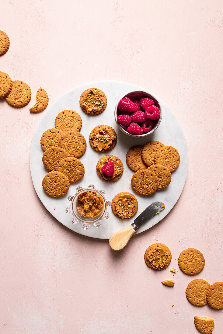 Draufsicht auf Teller mit köstlichen, knusprigen, hausgemachten Keksen und frischen Himbeeren, die auf einem Tisch mit einer Teekannenschale mit Samen serviert werden