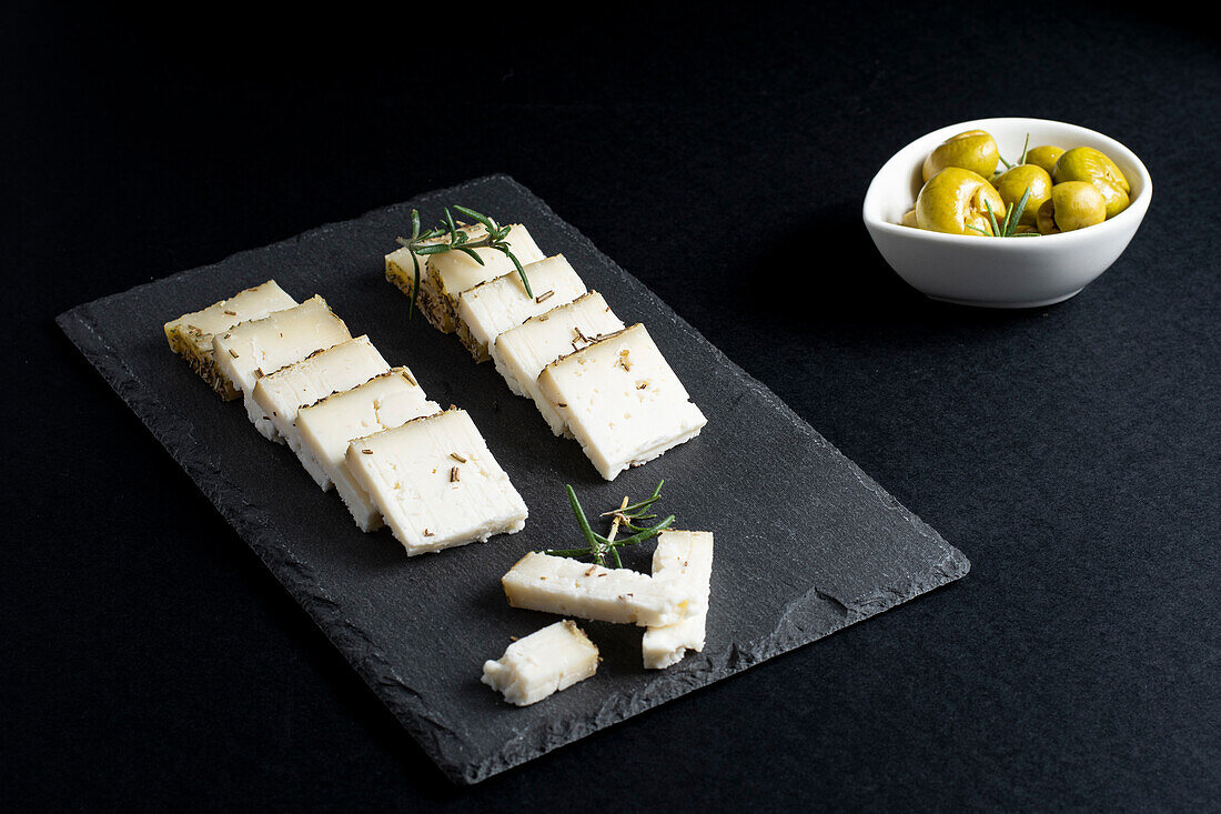 Appetitanregender frischer Schnittkäse auf einem Brett mit Rosmarinzweigen vor schwarzem Hintergrund