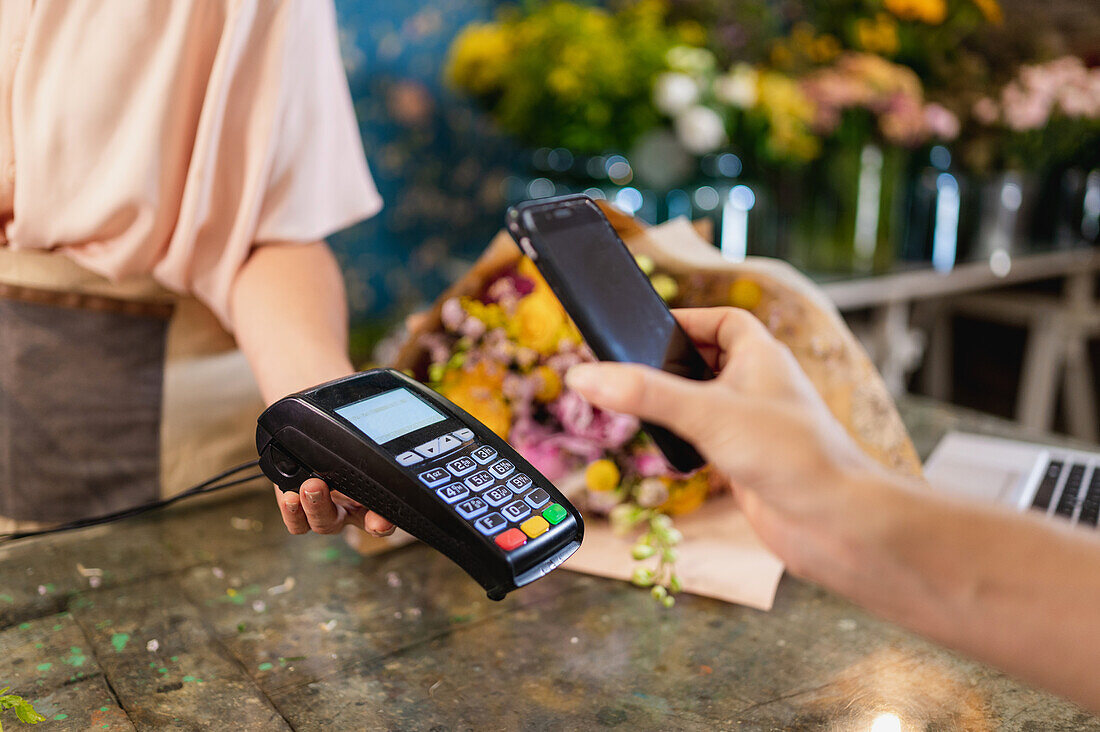 Crop unrecognizable client making contactless payment with mobile phone on POS terminal in floral shop