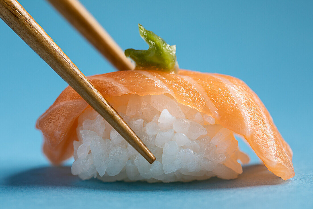 Leckeres japanisches Nigiri mit frischem Lachs und grünem Wasabi vor blauem Hintergrund mit Stäbchen in einem hellen Studio