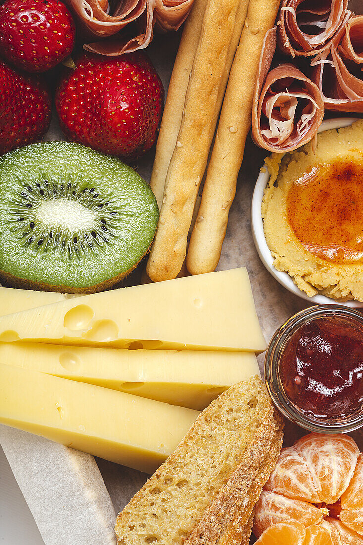 Von oben Nahaufnahme einer Brunchbox mit verschiedenen Wurst- und Käsesorten und Knäckebrot in der Nähe von reifen Kiwis, süßen Erdbeeren und geschälten Mandarinen in der Nähe von Marmelade in einem Glasgefäß auf buntem Hintergrund