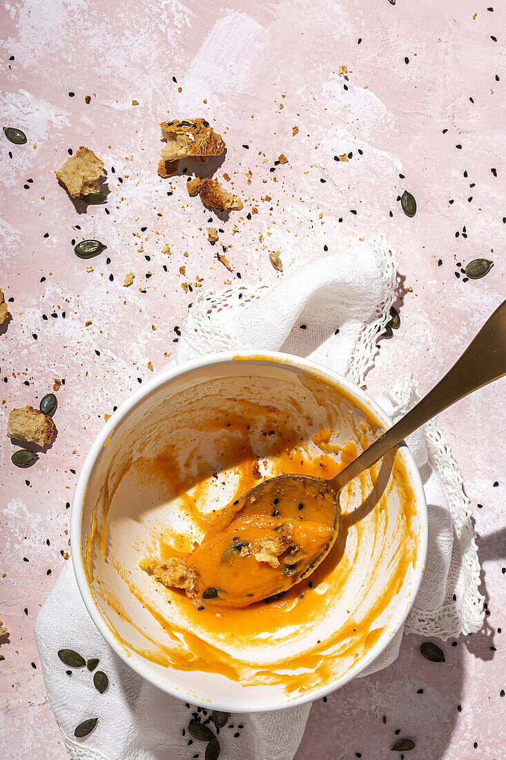 Top view of spoon in white ceramic bowl with eaten pumpkin cream soup placed on table near scattered seeds and crispy bread pieces