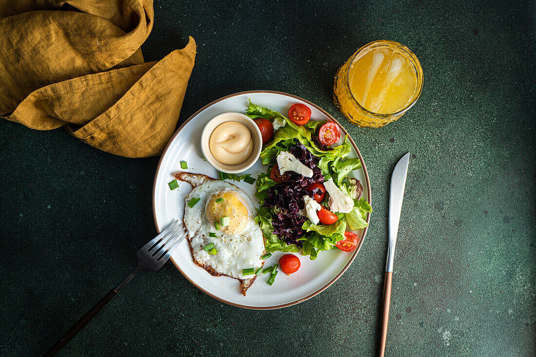 Traditionelles Mittagessen mit frischem Gemüsesalat, Spiegelei und Würstchen, serviert auf einem grünen Betontisch mit einem Glas frisch gepressten Orangensaft