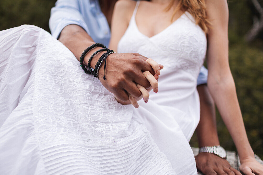 Crop unrecognizable ethnic groom with bracelets on wrist holding hand of bride in white wedding dress
