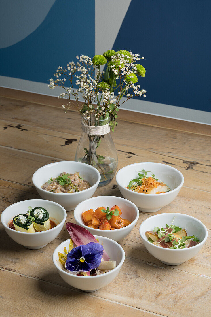 High angle of assorted tasty dishes in ceramic bowls placed on wooden floor near glass vase with fresh blooming flowers