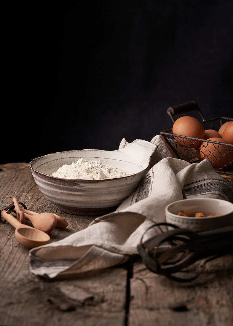 Heap of eggs in basket and bowl of flour placed on wooden table with various spoons and whisk before baking