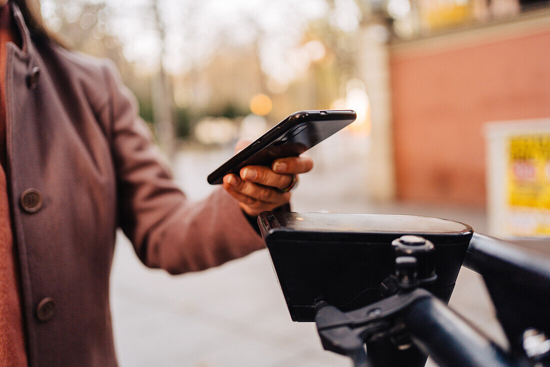 Selektiver Fokus einer anonymen Frau, die eine Smartphone-Anwendung verwendet und den Code eines Elektrorollers auf einer Straße in der Stadt scannt