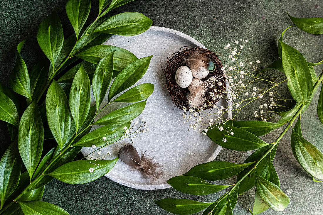 Von oben gedeckter Ostertisch mit Keramiktellern und Ostereiern in einem Nest, umgeben von italienischem Ruscus und Polsterblättern des Schleierkrauts