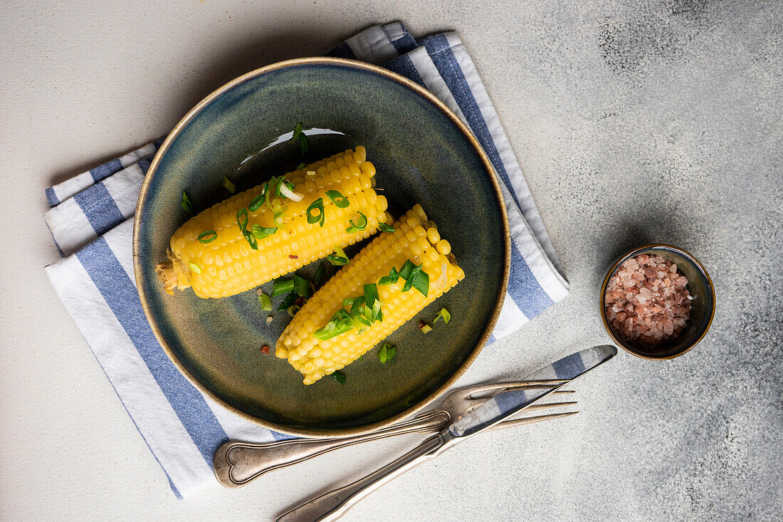 Top view of boiled corn with salt and green onion served on plate on grey background