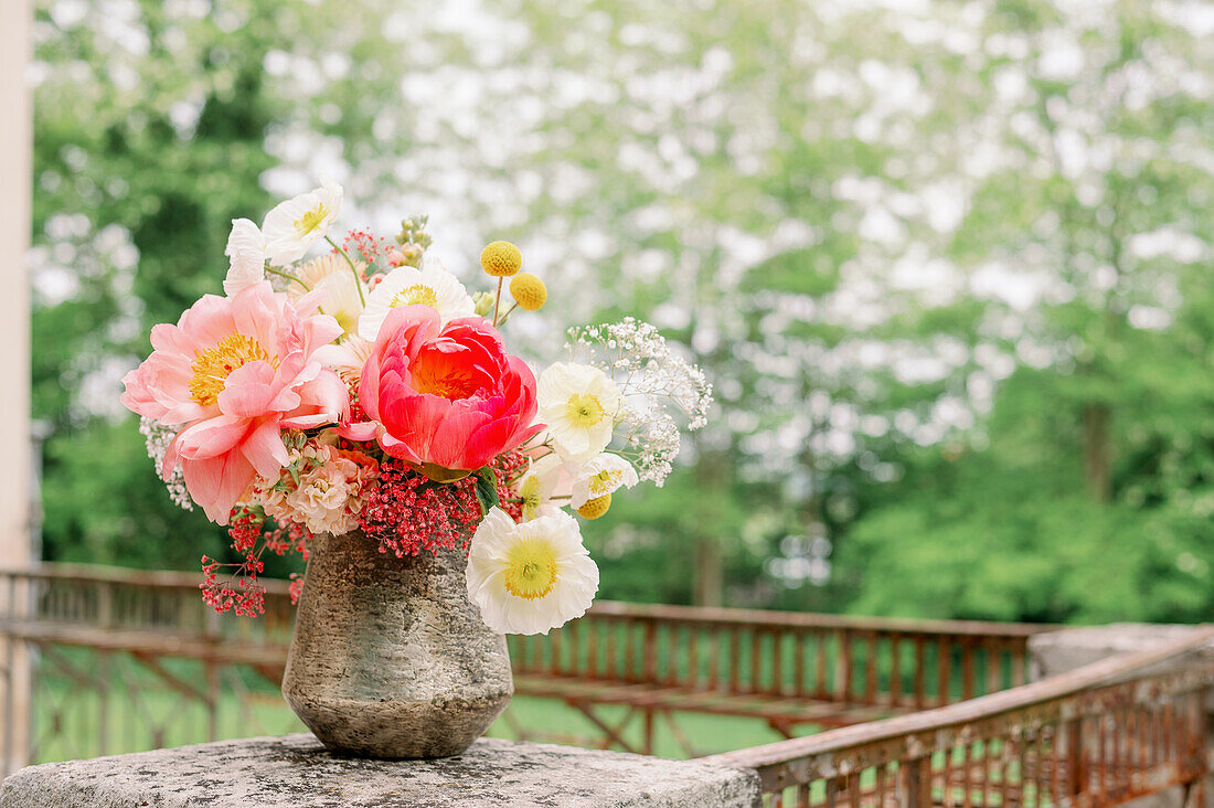 Shabby stone vase with bouquet of fresh peony and poppy flowers placed on terrace border on blurred background of lush trees in summer