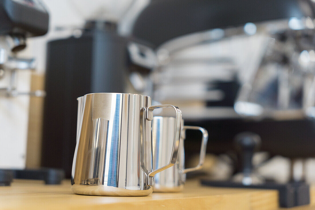 Soft focus of stainless professional pitchers for pouring milk placed on wooden counter in modern coffee house with coffeemaker machines