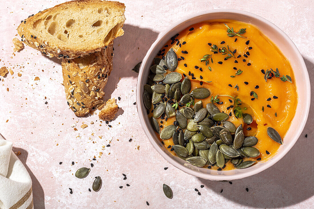 Draufsicht auf appetitliches hausgemachtes Kürbispüree mit Kernen und Kräutern in einer Schüssel auf dem Tisch neben knusprigen Brotscheiben in der Küche