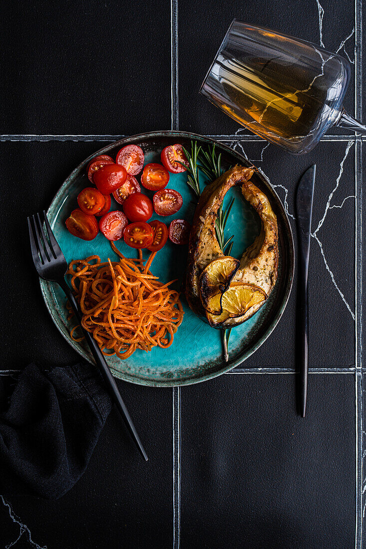 Lachssteak von oben mit würzigen Karotten und Kirschtomaten, serviert mit einem Glas trockenen Weißwein auf einem schwarz gekachelten Tisch