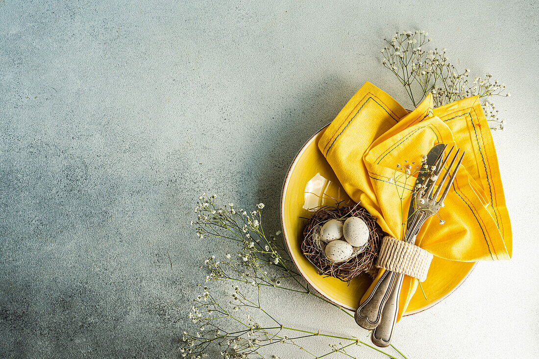 From above place setting for Easter dinner with yellow ceramic plates near nest with easter eggs surrounded by cushion baby's-breath leaves