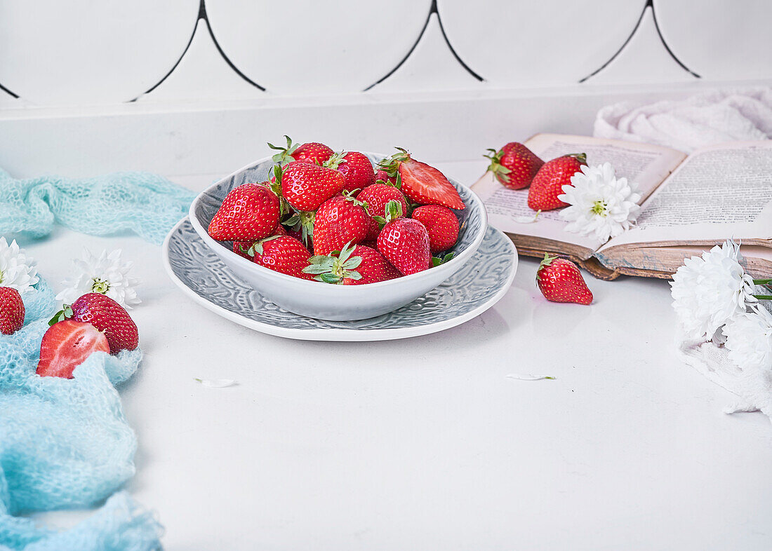 From above bowl of ripe strawberries placed on counter near fresh white flowers and opened book in daytime at home