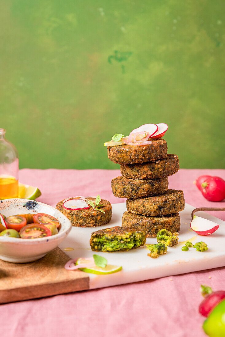 Vegan fritters stack served on cutting board near bowl of cherry tomatoes against green background