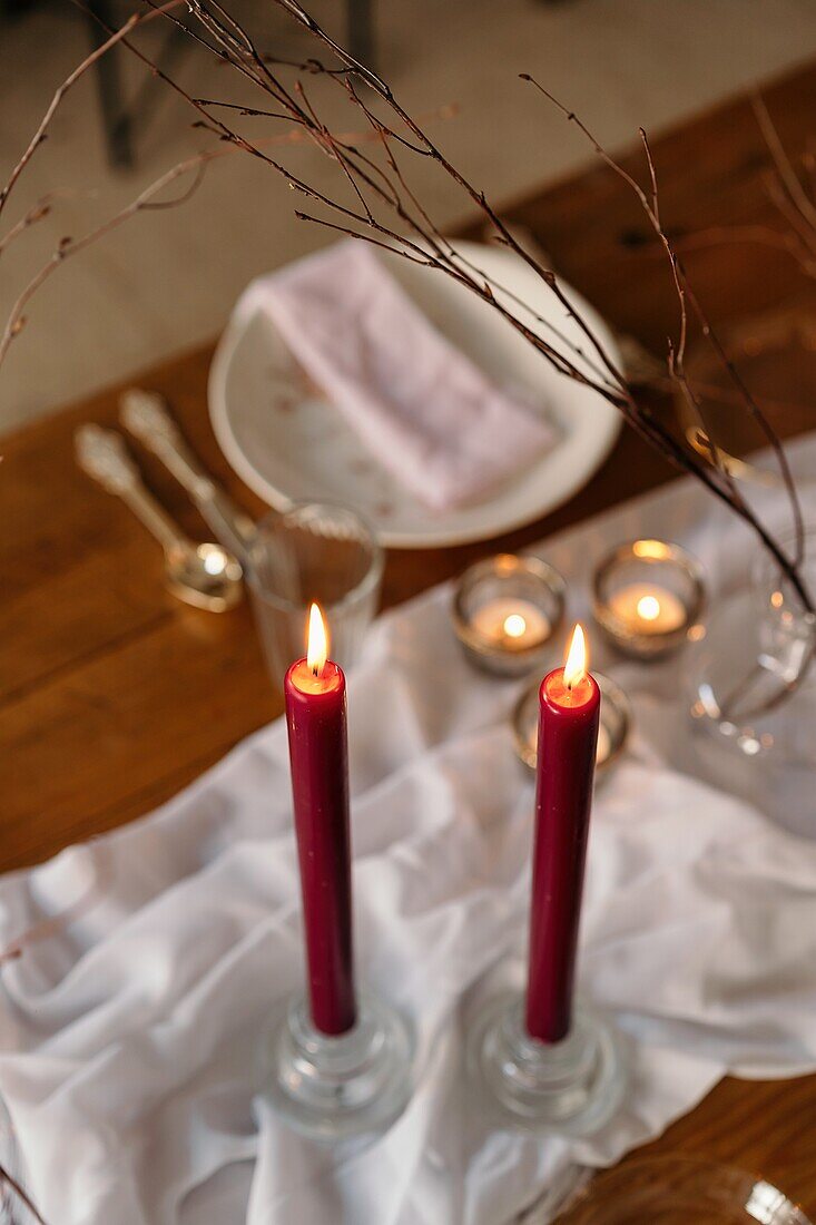 Weißes Tischtuch und Teller auf einem festlich gedeckten Tisch mit brennenden Kerzen und trockenen Baumzweigen von oben