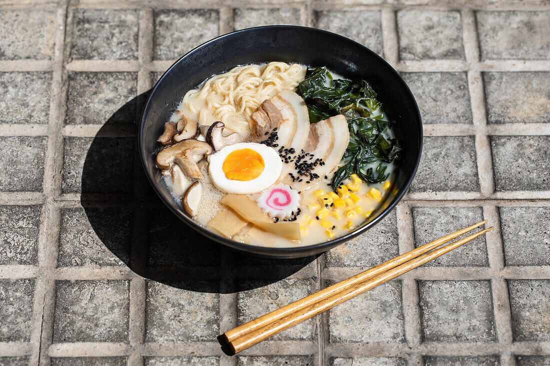 Von oben auf appetitliche japanische Ramen mit Pilzen und Ei, die auf dem gepflasterten Bürgersteig mit Stäbchen auf einer sonnigen Straße in der Stadt serviert werden