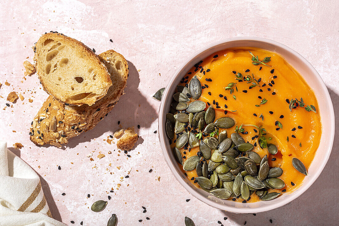 From above of appetizing homemade pumpkin puree with seeds and herbs in bowl placed on table near crispy bread slices in kitchen