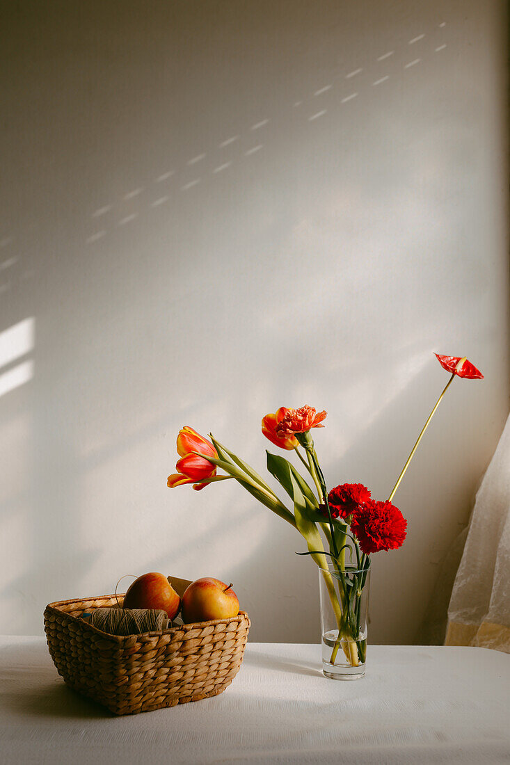 Vase mit blühenden Tulpen und Nelken auf dem Tisch neben Äpfeln in einer Weidenschale