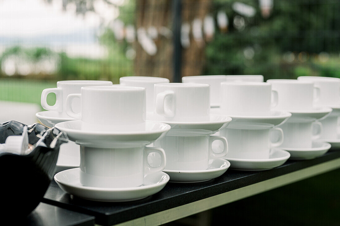 Pile of white ceramic cups with saucers arranged on counter near bowl with mini coffee creamers in outdoor cafe