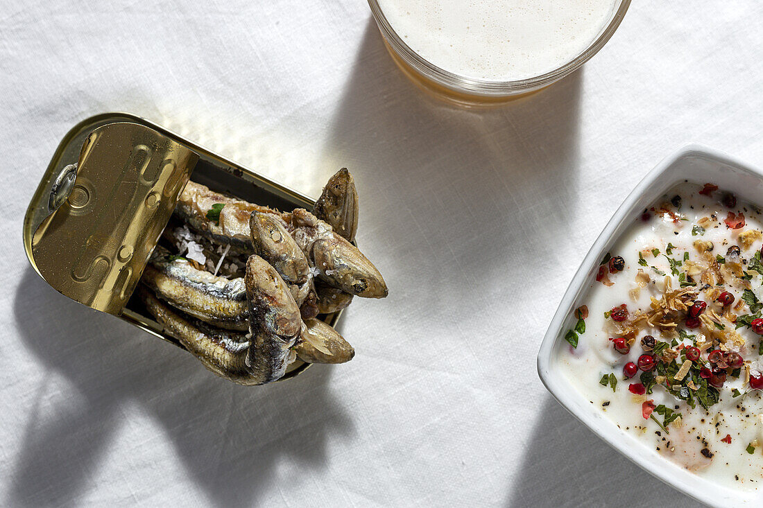 Draufsicht auf Sardellen in Dosen, serviert auf einem weißen Tisch mit einem Glas schäumendem Bier und weißer Gazpacho-Suppe